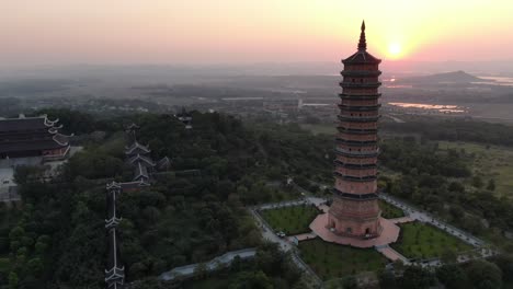 Drone-aerial-view-in-Vietnam-flying-over-a-buddhist-temple-area-filled-with-green-trees-and-a-pagoda-in-front-of-the-sun-in-Ninh-Binh-at-sunset