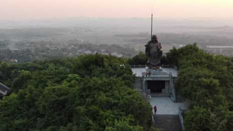 Drohnen-Luftaufnahme-In-Vietnam,-Die-Bei-Sonnenuntergang-Auf-Eine-Von-Tempeln-Umgebene-Buddha-Statue-In-Einem-Mit-Grünen-Bäumen-Bedeckten-Tal-In-Ninh-Binh-Zufliegt