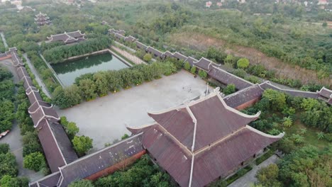 Drone-aerial-view-in-Vietnam-flying-over-a-buddhist-temple-area-filled-with-green-trees-in-front-of-a-serpent-river-in-Ninh-Binh-at-sunset