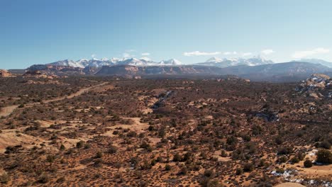 A-high-flying-drone-shot-over-a-remote-dirt-road-cutting-through-the-vast-and-unique-desert-land-near-Moab,-Utah,-with-the-snowy-Rocky-Mountains-towering-in-the-distance