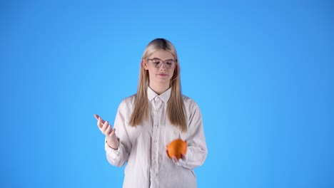 Junge-Blonde-Kaukasische-Smart-Dame-Mit-Brille-Spielt-Mit-Einem-Orangefarbenen-Obst-Studio-Unendlich-Chroma-Hintergrund-Torso-Schuss