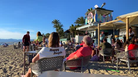 People-sitting,-talking-and-enjoying-life-at-a-beach-restaurant-in-Marbella,-woman-wearing-a-pullover-with-happy-people-words,-sunny-summer-day-with-blue-sky-in-Spain,-4K-shot