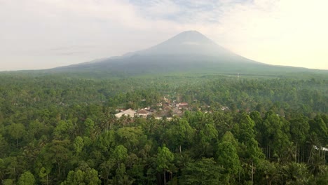 An-enchanting-aerial-vista-reveals-a-quaint-village-embraced-by-verdant-forest,-while-a-majestic-volcano-looms-gracefully-in-the-background,-a-timeless-spectacle-of-nature's-harmony