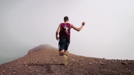 Eine-Aus-Der-Untersicht-Aufgenommene-Verfolgerkamera-Zeigt-Einen-Schwarzhaarigen-Mann-In-Shorts-Und-Tanktop,-Der-Auf-Einem-Isolierten,-Wolkenbedeckten-Felsigen-Bergkamm-Läuft