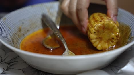 Corn-Cob-Being-Dipped-In-Spice-Sauce-Inside-Bowl