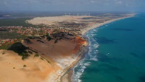 Aerial-view-of-the-sea,-waves,-cliffs,-a-small-village-and-a-wind-energy-at-background,-Morro-Branco,-Ceara,-Brazil