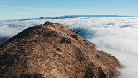 Pico-De-Montaña-Que-Se-Eleva-Sobre-Un-Mar-De-Nubes-Bajo-Un-Cielo-Azul-Claro,-Amplia-Toma-Aérea