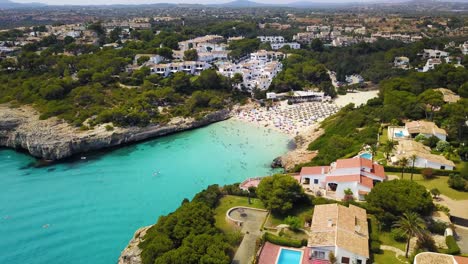 Cala-anguila-with-crystal-clear-waters,-beachgoers,-and-surrounding-villas,-sunny,-aerial-view