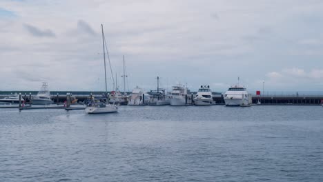 A-small-white-sailing-boat-has-entered-the-marina-and-is-preparing-to-dock,-with-other-larger-boats-moored-nearby