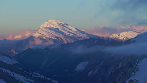 Captivating-aerial-perspective-offering-a-mesmerizing-view-of-sunrise-casting-a-warm-glow-on-the-silhouette-of-a-snowy-mountain-range,-filmed-by-drone