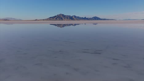 Drone-flying-at-low-altitude-over-water-surface-of-flooded-Bonneville-Salt-Flats-with-rocky-mountain-in-background,-Utah