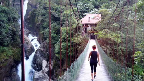 Woman-walking-towards-a-cabin-over-a-suspension-bridge-in-Baños,-Ecuador,-surrounded-by-lush-greenery-and-rushing-river-below