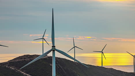 Closeup-of-Rotating-windmills-above-sunset-skyline-and-mountains-time-lapse-sun-background,-motion-of-renewable-energy-fans-in-countryside-landscape