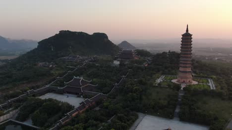 Drone-aerial-view-in-Vietnam-flying-away-from-a-buddhist-temple-and-pagoda-area-in-Ninh-Binh-at-sunset-surrounded-by-green-mountains