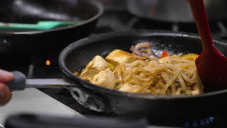 Spatula-stirs-sizzling-flat-rice-noodle-tofu-dish-over-hot-flame-in-commercial-restaurant-kitchen,-chef-shakes-and-tosses-vegetarian-pad-kee-mao,-slow-motion-close-up-4K