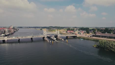 Puente-Elevado-Sobre-Un-Río-Tranquilo-Con-Barcos-En-Movimiento,-Casas-Al-Fondo,-Día-Soleado,-Vista-Aérea