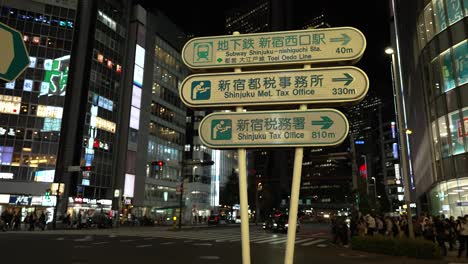 A-street-sign-in-Tokyo's-bustling-Shinjuku-district-at-night,-amidst-the-flow-of-traffic-and-pedestrians-crossing-the-road,-epitomizing-the-concept-of-urban-dynamism-and-nocturnal-activity