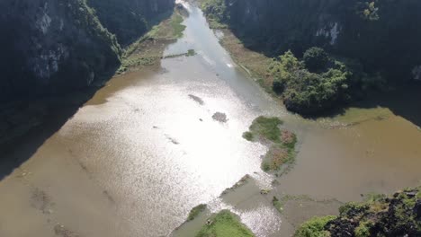 Drone-aerial-view-in-Vietnam-vertical-panning-showing-rocky-mountains-covered-with-green-trees-over-a-wide-brown-river-in-Ninh-Binh-on-a-sunny-day