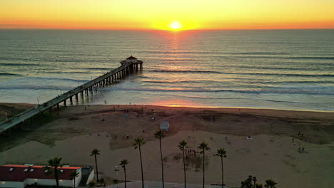 Ein-Ruhiger-Sonnenuntergang-über-Einem-Pier-Mit-Palmen-Und-Ruhigem-Meer,-Die-Aktivitäten-Am-Strand-Gehen-Zu-Ende,-Luftaufnahme