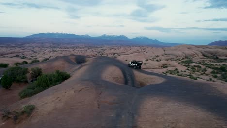 Eine-4K-Drohnenaufnahme-Eines-Hochgefahrenen-Jeeps,-Der-Im-Gelände-Durch-Die-Extreme-Und-Felsige-Wüstenlandschaft-In-Der-Nähe-Von-Moab,-Utah,-Fährt,-Mit-Den-Schneebedeckten-Rocky-Mountains-In-Der-Ferne