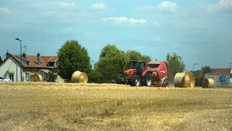Red-tractor-with-large-bundling-machine-collecting-dry-yellow-grass-of-field