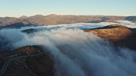 Clouds-rolling-over-mountainous-landscape-at-dawn,-road-winding-through-hills,-aerial-view