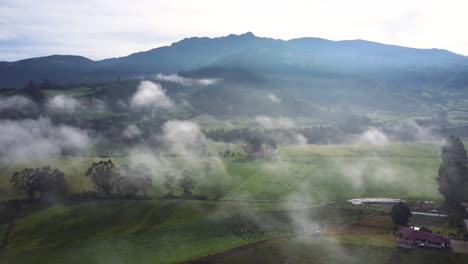 Dron-Aéreo-De-Movimiento-Lento-Hacia-Atrás-Sobre-Los-Campos-Y-Las-Nubes-Con-El-Volcán-Pasochoa-Al-Fondo-En-Neblina,-Ecuador