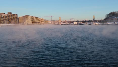Vuelo-Bajo-Aéreo-Sobre-Aguas-Con-Niebla-Flotante-Que-Se-Eleva-Para-Revelar-Bjorvika-En-El-Distrito-Centrum-De-Oslo,-Noruega