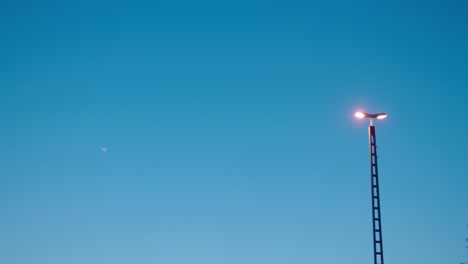 Straßenlaterne-Leuchtet-Gegen-Den-Abendhimmel,-Mit-Einem-Schwachen-Mond-Sichtbar