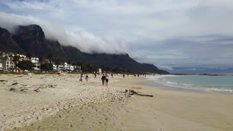 People-and-dogs-walk-on-Camp's-Bay-Beach-under-cloud-covered-hills