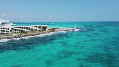 Cancun-coastline-with-lighthouse-and-hotel,-clear-blue-waters-under-sunny-skies,-aerial-view