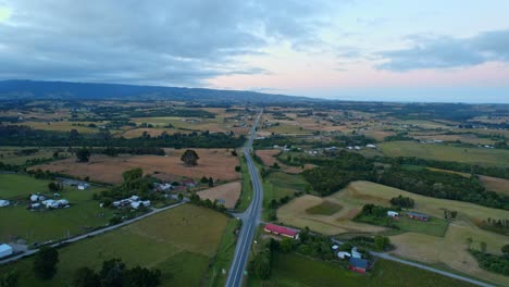 Chiloé,-Chile-Al-Atardecer-Con-Un-Camino-Sinuoso-En-Medio-De-Campos-Verdes-Y-Casas-Dispersas,-Vista-Aérea