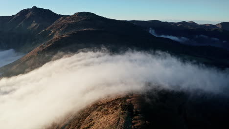 Misty-mountains-with-sunlit-peaks-and-valleys,-clouds-rolling-over-the-landscape-in-an-aerial-shot