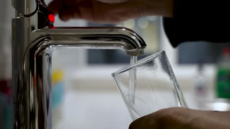 Primer-Plano-De-Una-Mano-Llenando-Un-Vaso-Transparente-Con-Agua-De-Un-Grifo-Moderno,-Ambiente-De-Cocina