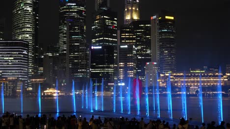 Magische-Licht--Und-Wassershow-Von-Spectra-Mit-Menschen,-Die-Sich-Auf-Der-Marina-Bay-Sands-Event-Plaza-Versammelt-Haben,-Im-Hintergrund-Die-Innenstadtansicht,-Im-Zeitraffer-Eine-Statische-Aufnahme-Des-Lebhaften-Nachtunterhaltungsprogramms-In-Singapur