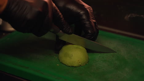 Chef-hands-wearing-black-latex-gloves-slicing-soft-green-avocado-half-on-chopping-board