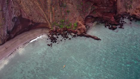 Vista-Aérea-Cinematográfica-Que-Captura-A-Turistas-Remando-En-Un-Colorido-Kayak-En-Un-Exótico-Destino-Tropical-Hawaiano-Con-Una-Tranquila-Superficie-Turquesa-Del-Océano-Y-Acantilados-De-Tierra-Roja.