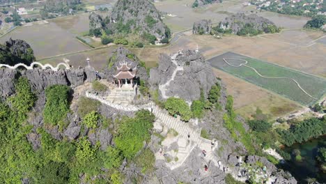 Drone-aerial-view-in-Vietnam-flying-down-in-front-of-rocky-mountain-covered-with-green-trees-with-stairs-and-a-temple-in-Ninh-Binh-on-a-sunny-day