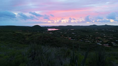 Drone-ascend-above-cactus-on-hills-to-reveal-Curacao-countryside,-fire-red-glow-in-wispy-sunset-clouds