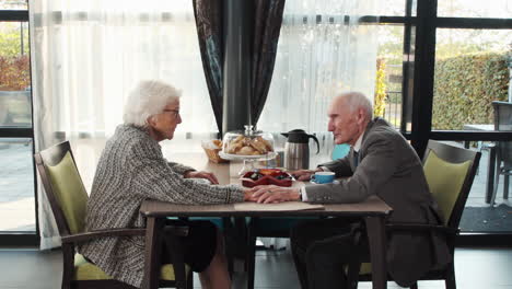 Lovable-elderly-couple-sitting-at-table-reaching-out-and-holding-hands