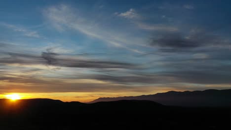 Flug-In-Einen-Sonnenuntergang-In-Einem-Tal-Mit-Einem-Gebirgssystem-Von-Dunkler,-Fast-Schwarzer-Farbe-Mit-Einem-Gelben,-Blauen-Himmel-Und-Grauen-Wolken-Und-Dem-Sonnenuntergang-In-Den-Bergen,-Den-Man-Mit-Einer-Wendung-In-Spanien-Beobachtet