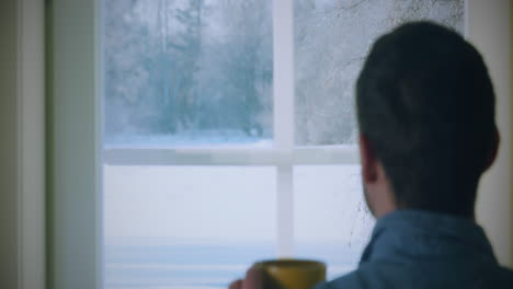 Man-in-blue-shirt-enjoying-hot-drink,-gazing-through-frosty-window,-feeling-cozy-indoors