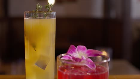 Nahaufnahme-Eines-Orangencocktails-Mit-Thymiangarnitur-Und-Eines-Roten-Cocktails-Mit-Blumengarnitur-In-Einem-Japanischen-Sushi-Restaurant,-Wobei-Das-Video-In-Zeitlupe-Nach-Oben-Geschwenkt-Wird