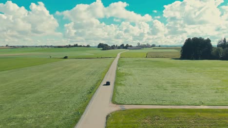 A-single-car-driving-on-a-long,-winding-road-through-vibrant-green-fields-under-a-partly-cloudy-sky,-daytime,-aerial-view