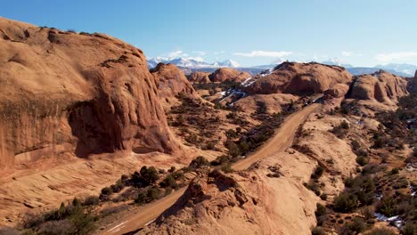 A-high-flying-drone-shot-over-a-remote-dirt-road-cutting-through-the-vast-and-unique-desert-land-near-Moab,-Utah,-with-the-snowy-Rocky-Mountains-towering-in-the-distance