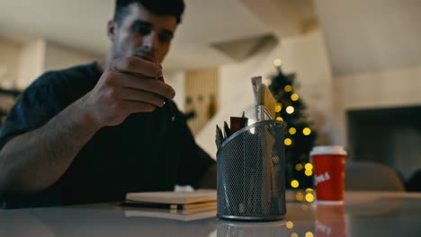 A-profile-shot-capturing-artist-as-he-selects-pencils-from-a-wire-mesh-holder,-set-against-the-backdrop-of-an-out-of-focus-Christmas-tree-and-bokeh-fairy-lights