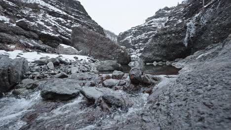 Icy-stream-flowing-through-a-snowy-mountain-ravine,-overcast-sky
