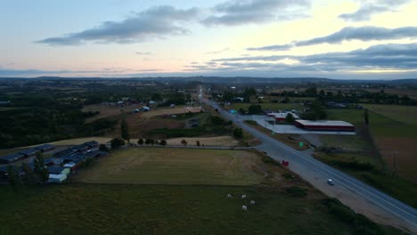 Aerial-orbit-of-a-meadow-with-a-lonely-road-in-Chiloé-with-an-orange-and-bluish-background-with-clouds,-Chile