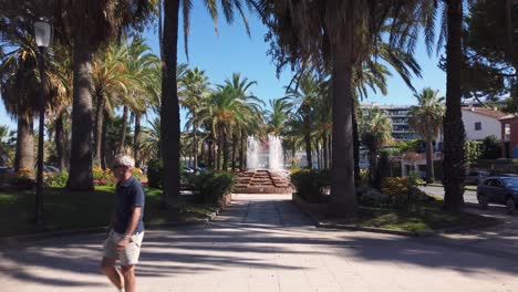 Man-in-sunglasses-walks-through-frame-in-park-with-fountain-in-Antibes