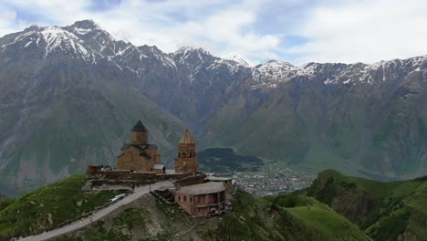 Drone-aerial-view-in-Georgia-flying-by-Gergeti-Trinity-medieval-orthodox-church-in-Kazbegi-surrounded-by-green-mountains-valley-with-snowed-peaks